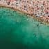 Aerial view of a crowded beach with swimmers and colorful umbrellas along the shoreline. - Olive Oil Times