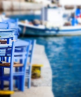 Set of blue wooden chairs and a table with a checkered tablecloth near a body of water. - Olive Oil Times