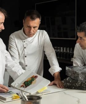 Three chefs in white jackets examining a cookbook while discussing culinary presentations. - Olive Oil Times