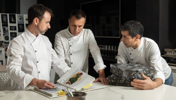 Three chefs in white jackets examining a cookbook while discussing culinary presentations. - Olive Oil Times