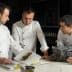 Three chefs in white jackets examining a cookbook while discussing culinary presentations. - Olive Oil Times