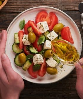 Hands pouring olive oil over a salad with tomatoes, cucumbers, olives, and feta cheese on a plate. - Olive Oil Times
