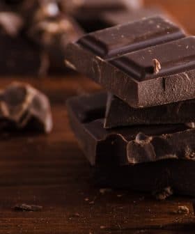 Several pieces of dark chocolate stacked on a wooden surface with additional chocolate fragments scattered around. - Olive Oil Times