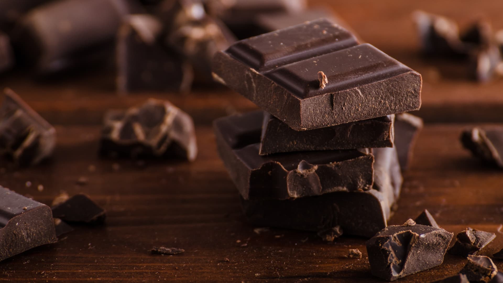 Several pieces of dark chocolate stacked on a wooden surface with additional chocolate fragments scattered around. - Olive Oil Times