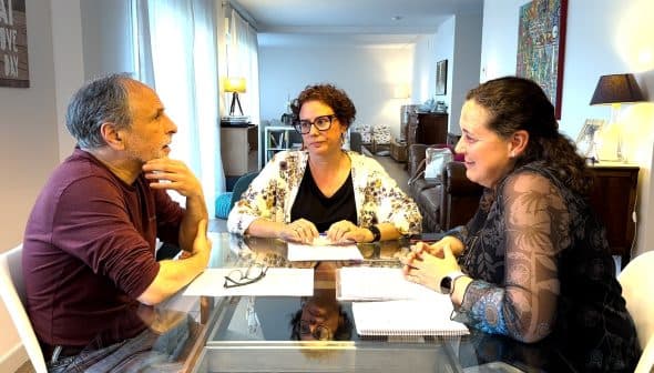 Three individuals seated at a table engaged in a discussion with papers in front of them. - Olive Oil Times