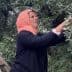 Woman wearing a headscarf picking olives from an olive tree while standing on a ladder. - Olive Oil Times