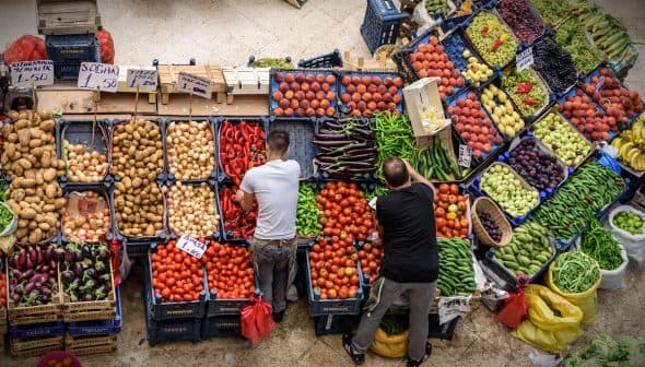 Two individuals arranging fresh vegetables and fruits at a market stall with various produce. - Olive Oil Times