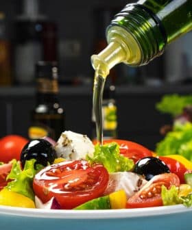 Olive oil being poured over a fresh salad with tomatoes, lettuce, and olives in a bowl. - Olive Oil Times