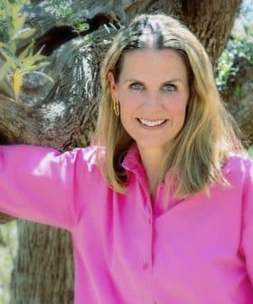 Woman with long hair wearing a pink shirt, leaning against an olive tree with a smile. - Olive Oil Times