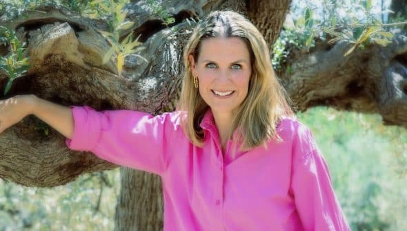 Woman with long hair wearing a pink shirt, leaning against an olive tree with a smile. - Olive Oil Times