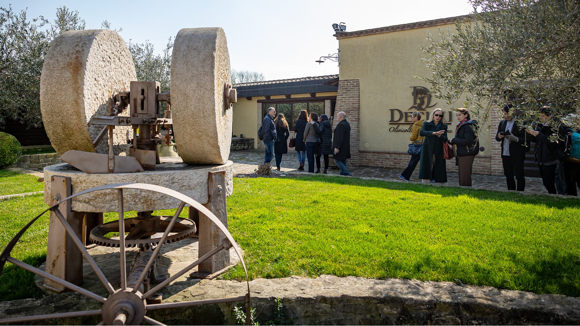Old stone olive oil pressing equipment displayed outdoors near a building with visitors observing. - Olive Oil Times