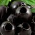 Close-up of black olives with a glossy surface, placed in a clear bowl with green leaves. - Olive Oil Times