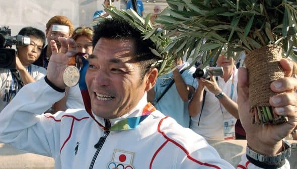 An athlete holding a medal and a laurel wreath while smiling at a crowd of photographers. - Olive Oil Times