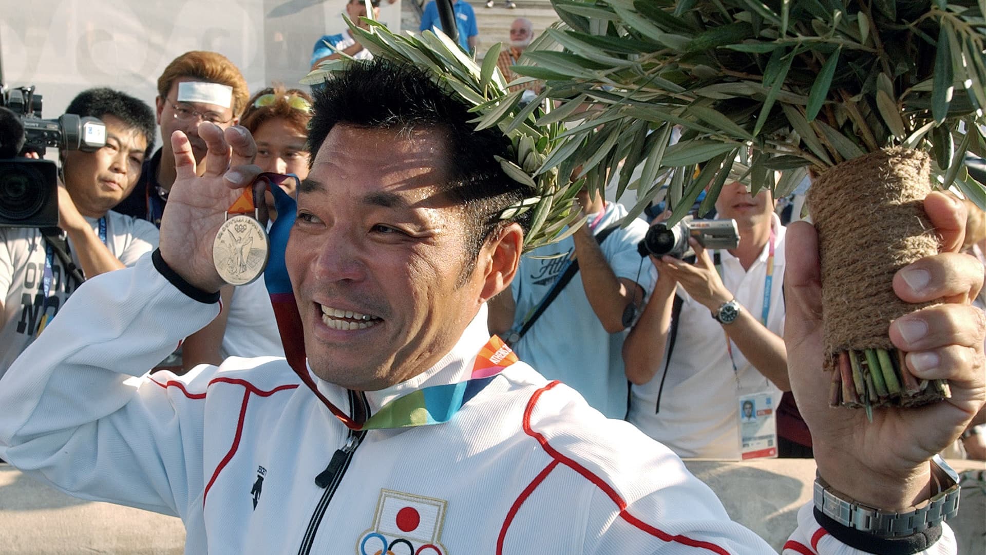 An athlete holding a medal and a laurel wreath while smiling at a crowd of photographers. - Olive Oil Times