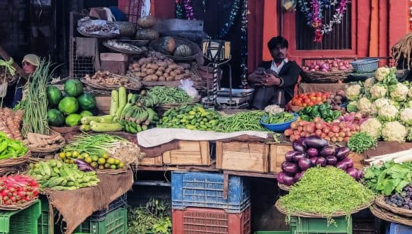 A variety of fresh vegetables displayed in baskets at a market, with a vendor seated nearby. - Olive Oil Times