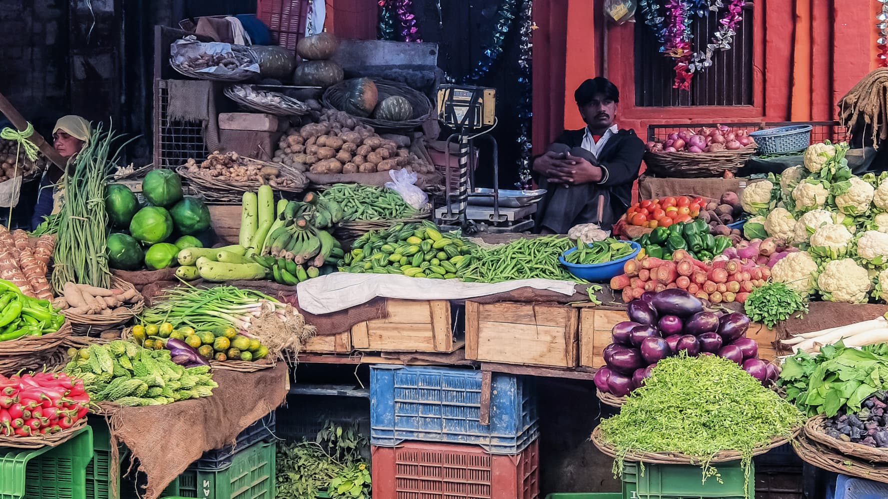 A variety of fresh vegetables displayed in baskets at a market, with a vendor seated nearby. - Olive Oil Times