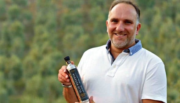 Man in a white polo shirt holding a bottle of olive oil with a blurred background. - Olive Oil Times