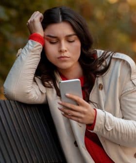 A woman with long dark hair wearing a light coat and red sweater, looking at her smartphone while sitting on a bench. - Olive Oil Times