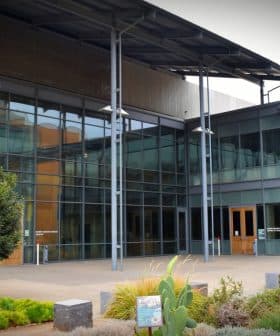 Entrance of a modern building featuring large glass windows and a metal roof structure. - Olive Oil Times