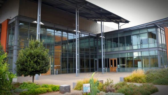 Entrance of a modern building featuring large glass windows and a metal roof structure. - Olive Oil Times