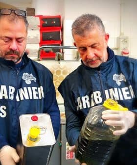 Two men in uniforms holding a metal container and a plastic bottle filled with oil. - Olive Oil Times