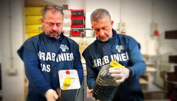 Two men in uniforms holding a metal container and a plastic bottle filled with oil. - Olive Oil Times