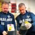 Two men in uniforms holding a metal container and a plastic bottle filled with oil. - Olive Oil Times