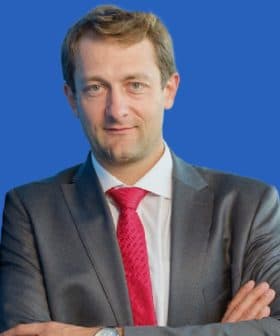 Man wearing a gray suit and a red tie, standing with arms crossed against a blue background. - Olive Oil Times