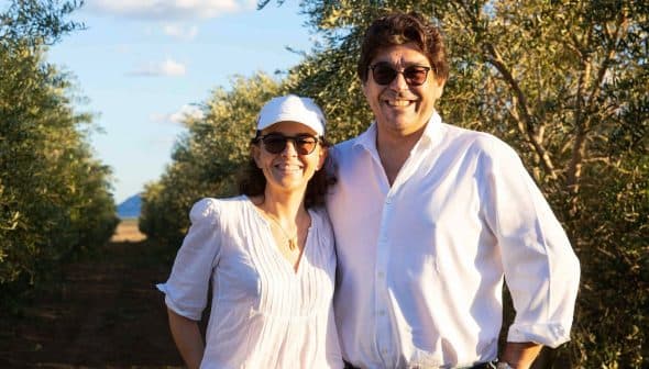 A man and woman smiling while standing in an olive grove under a clear sky. - Olive Oil Times