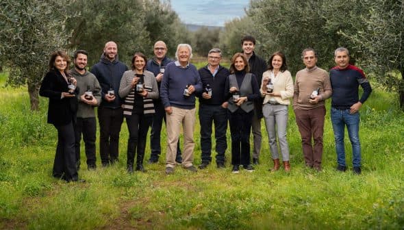Group of twelve individuals standing in an olive grove, each holding a bottle of olive oil. - Olive Oil Times