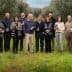 Group of twelve individuals standing in an olive grove, each holding a bottle of olive oil. - Olive Oil Times