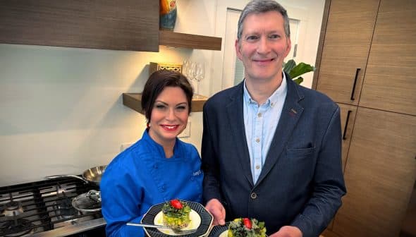 Two individuals, one in a blue chef jacket and the other in a blazer, holding plates of food in a kitchen. - Olive Oil Times