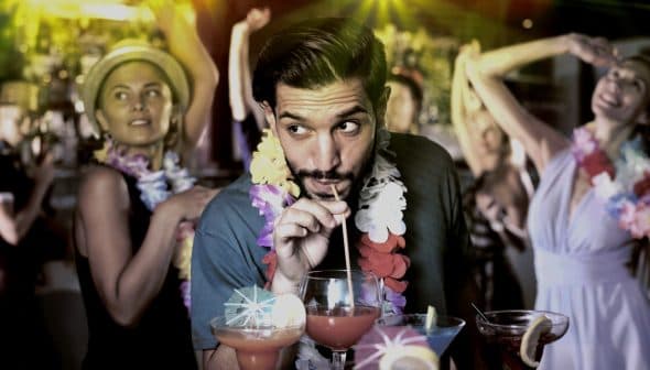 Man sipping a cocktail through a straw while surrounded by people at a party. - Olive Oil Times