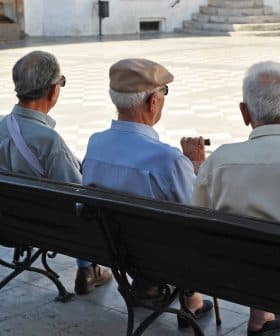 Three elderly men sitting on a bench, viewed from behind, in a public square. - Olive Oil Times