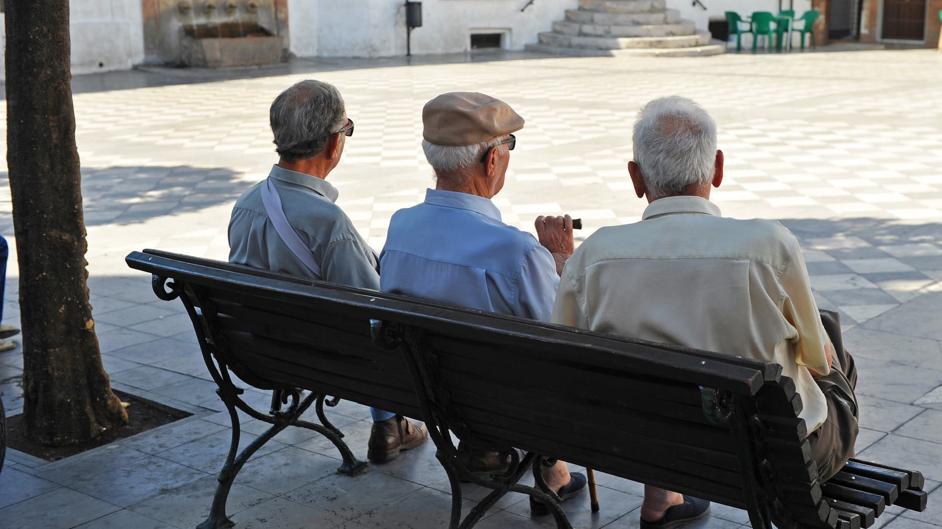 Three elderly men sitting on a bench, viewed from behind, in a public square. - Olive Oil Times