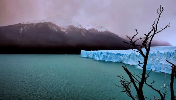 A glacier with a blue ice edge in a body of water, surrounded by mountains and a cloudy sky. - Olive Oil Times