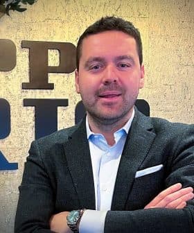 Man in a dark blazer and white shirt standing with arms crossed in front of a textured wall. - Olive Oil Times