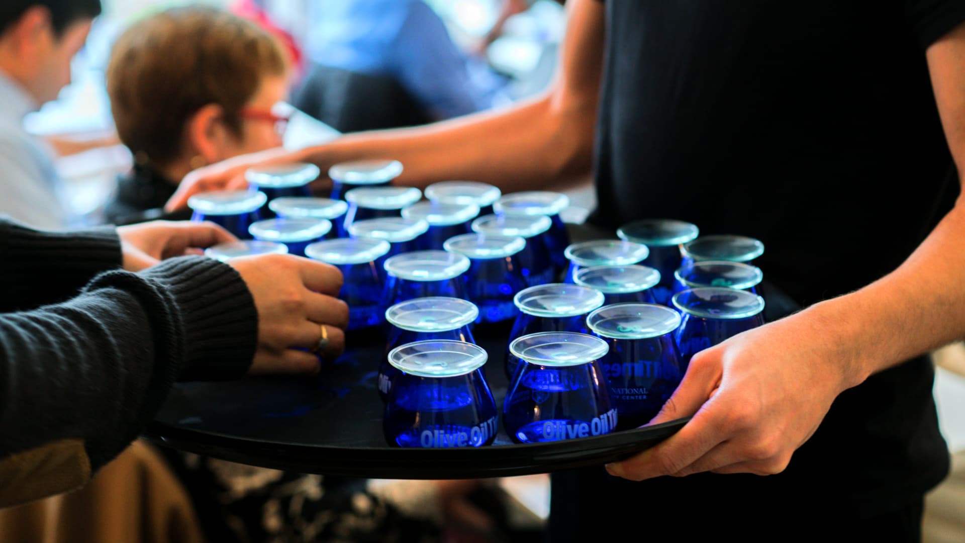A tray holding multiple blue glasses filled with olive oil being served at an event. - Olive Oil Times