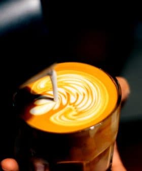 A glass of latte with intricate milk foam art on top, held in a hand against a dark background. - Olive Oil Times