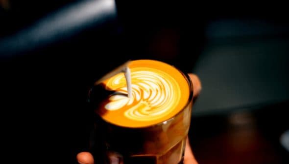 A glass of latte with intricate milk foam art on top, held in a hand against a dark background. - Olive Oil Times