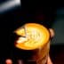 A glass of latte with intricate milk foam art on top, held in a hand against a dark background. - Olive Oil Times