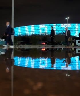 Group of people walking past a modern building with illuminated blue facade at night, reflected in a puddle. - Olive Oil Times