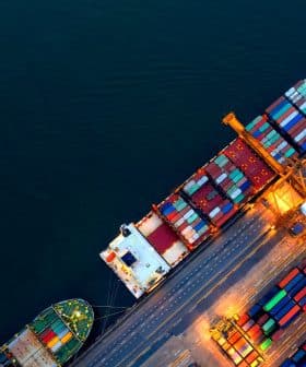 Aerial view of container ships docked at a port with colorful shipping containers stacked on the deck. - Olive Oil Times