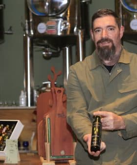 Man with a beard holding a bottle of olive oil in a production setting with equipment in the background. - Olive Oil Times