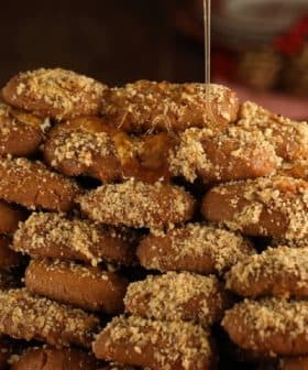 A large stack of honey-dipped cookies topped with crushed nuts on a wooden surface. - Olive Oil Times