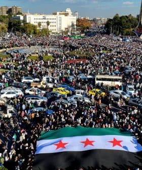 A large crowd gathered in a public square holding a Syrian flag with three stars. - Olive Oil Times