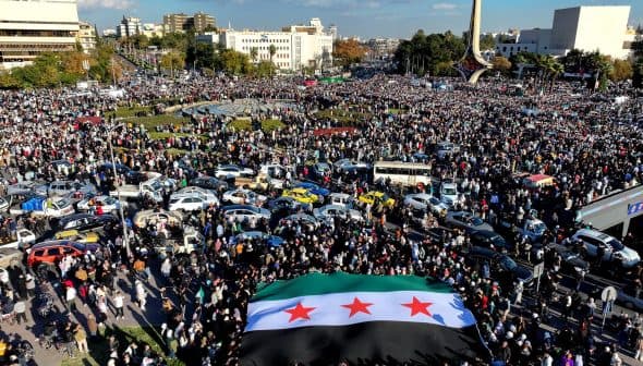 A large crowd gathered in a public square holding a Syrian flag with three stars. - Olive Oil Times