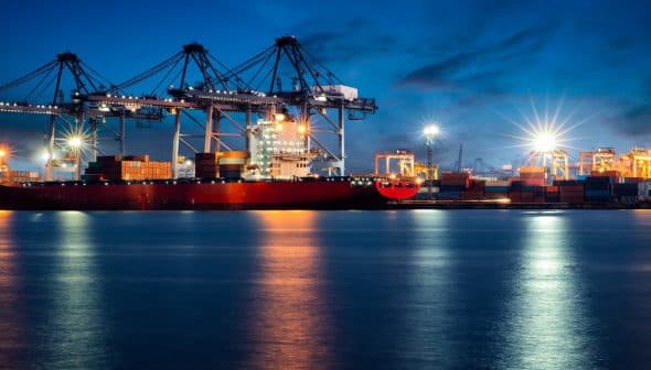 Large container ship docked at a port with cranes and cargo containers under a night sky. - Olive Oil Times