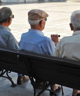 Three elderly men sitting on a park bench, viewed from behind, wearing hats and light-colored shirts. - Olive Oil Times