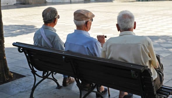 Three elderly men sitting on a park bench, viewed from behind, wearing hats and light-colored shirts. - Olive Oil Times